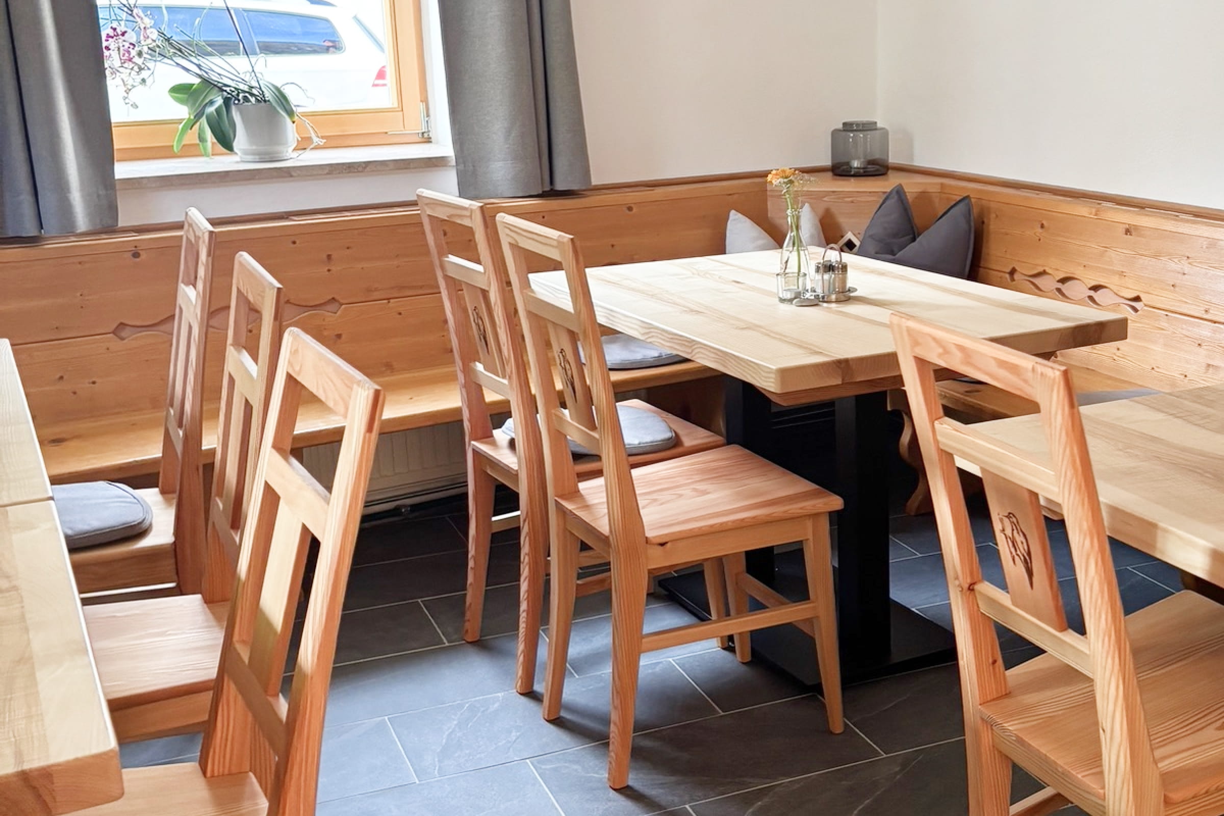 Moderner Gasthausinnenraum mit hellen Holztischen, Holzstühlen und einer Holzbank an der Wand unter einem Fenster mit grauem Vorhang