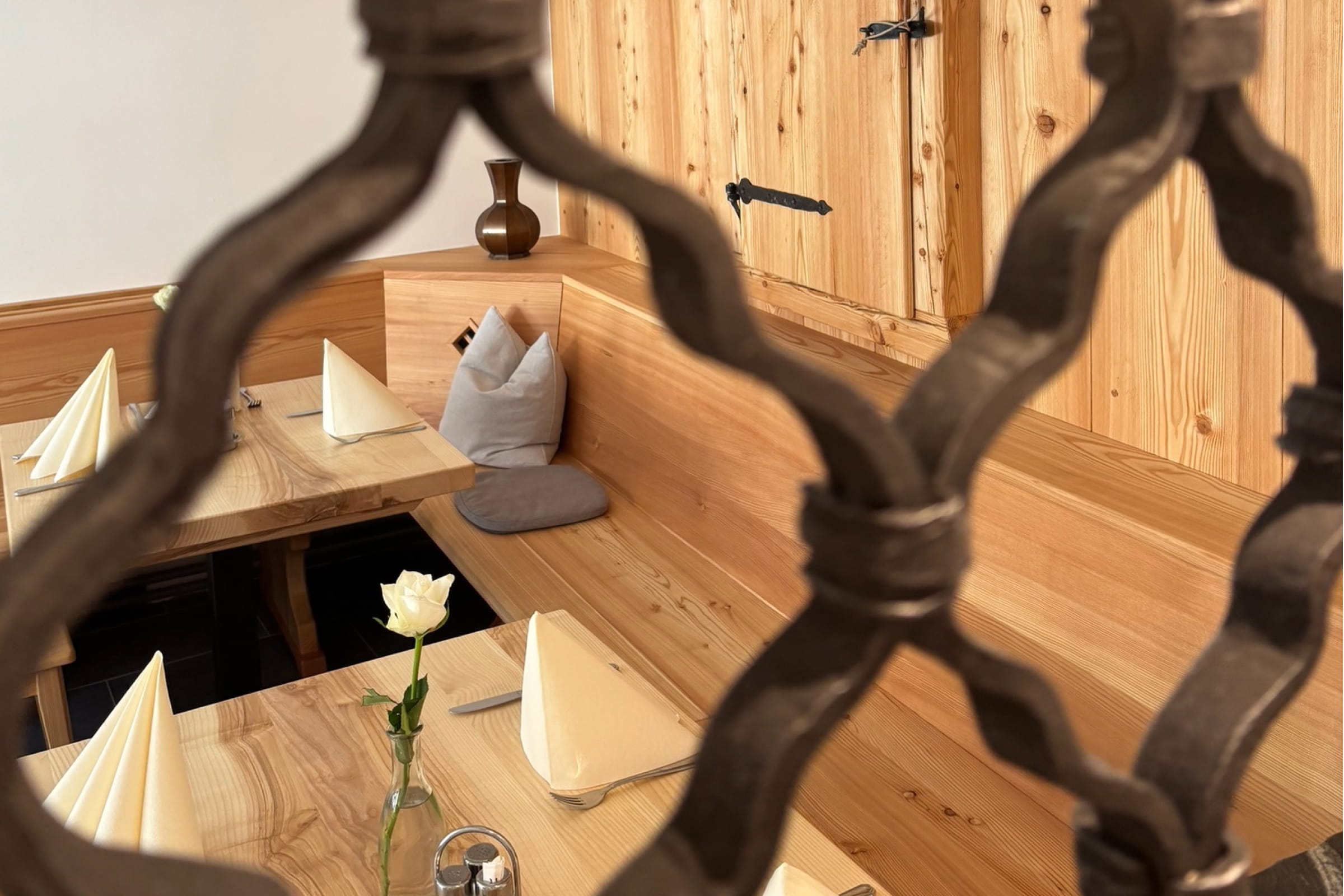 Blick durch ein dekoratives Gitter auf eine traditionelle Holzeckbank mit eingedeckten Tischen und einer weißen Rose in einer Vase