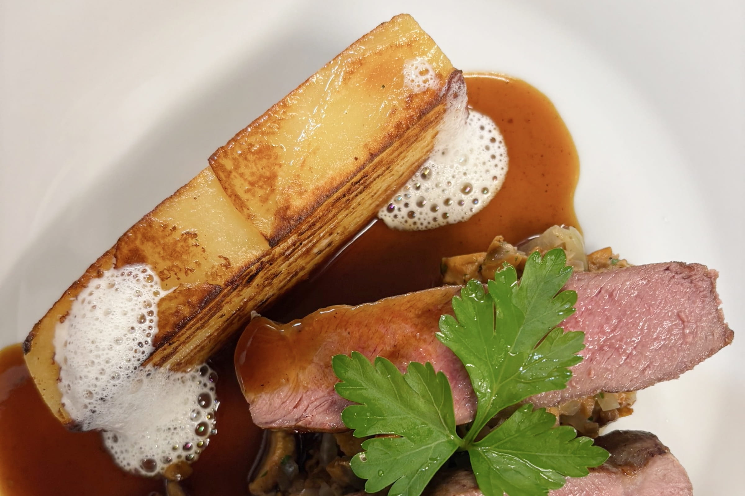 Anrichte mit rosa gebratenem Lammfleisch, rechteckigem Kartoffelgratin, brauner Sauce, Schaum und Petersilienblatt auf weißem Teller.