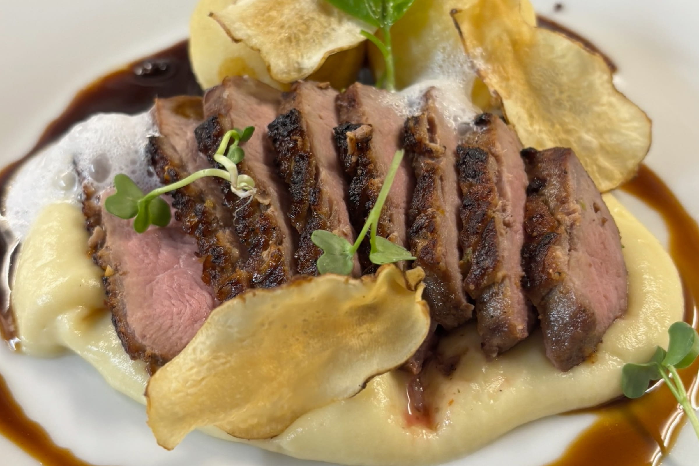 Auf einem Teller angerichtete Scheiben von rosa gebratenem Rindfleisch auf cremigem Püree mit brauner Sauce, garniert mit grünen Kräutern und knusprigen Chips