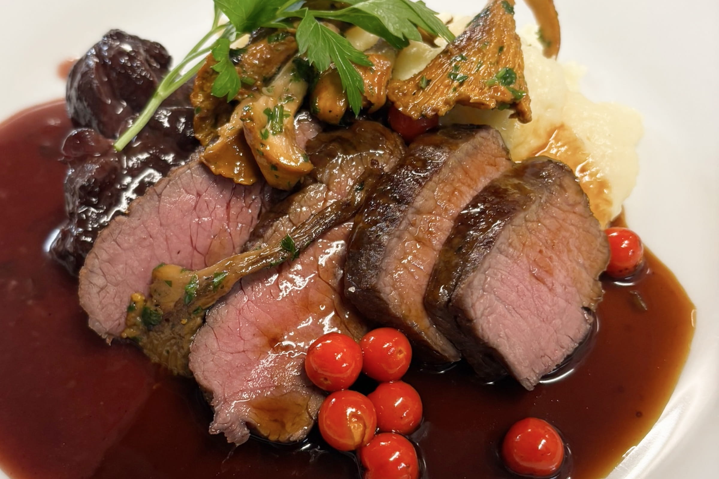 Drei rosa gebratene Rindfleischscheiben mit brauner Sauce, Pfifferlingen, kleinen roten Beeren und Petersilienblatt auf weißem Teller.
