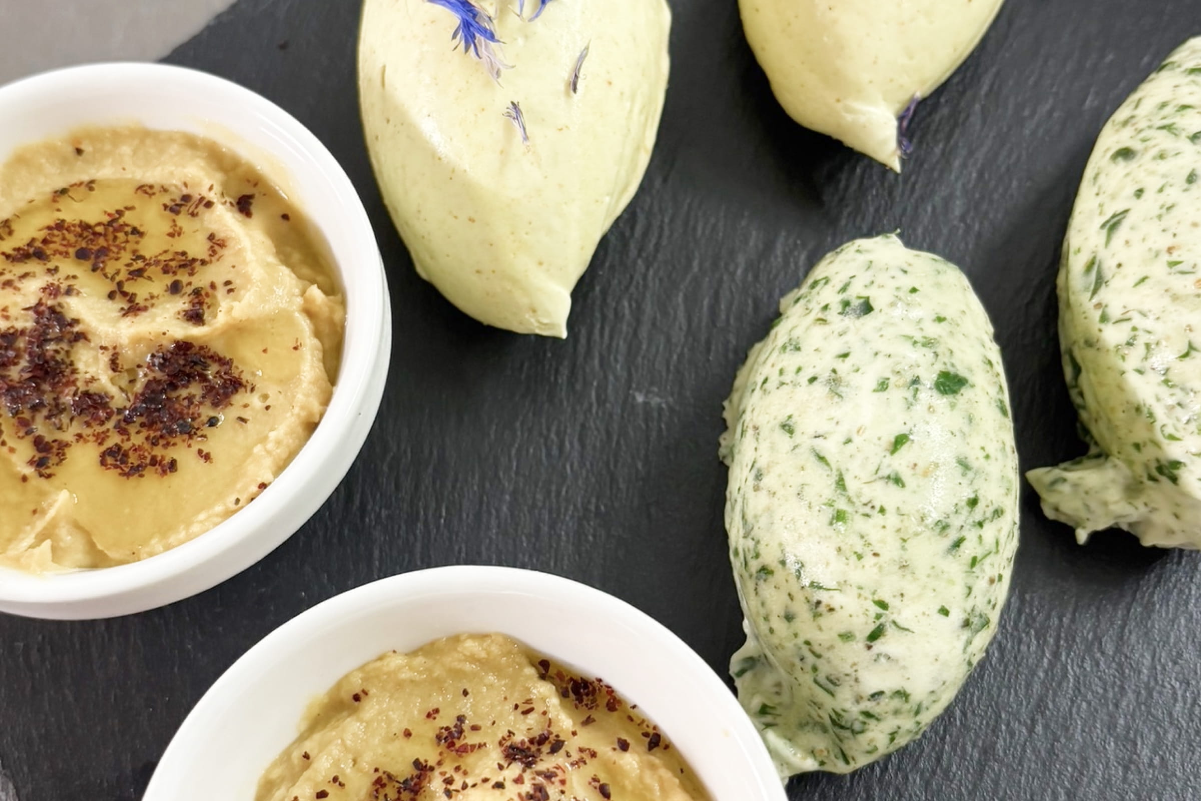 Mehrere Nocken verschiedener hausgemachter Dips auf dunkler Schieferplatte, darunter ein heller mit Kräutern und ein brauner mit Gewürzbestreuung.