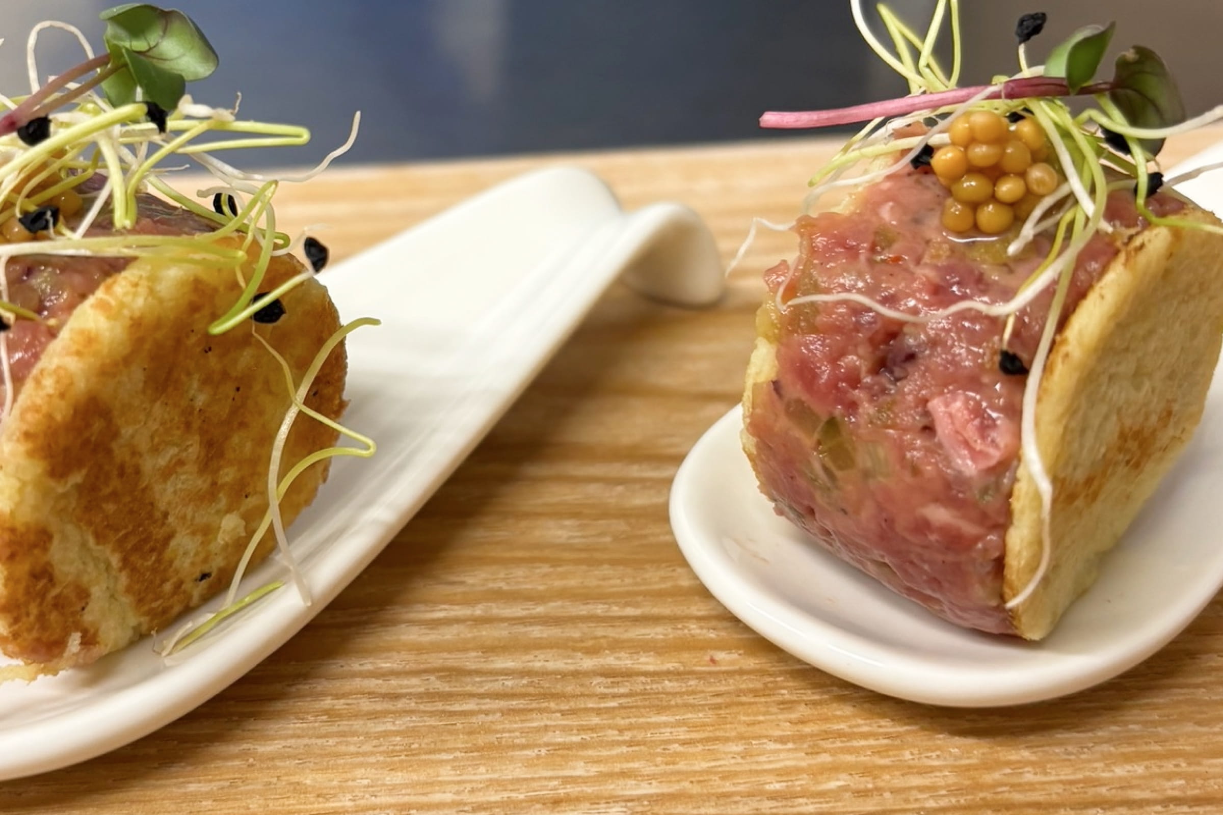 Zwei kleine runde Tartare-Portionen auf weißen Löffeln mit knusprigen Chips und Sprossen garniert auf Holzuntergrund.