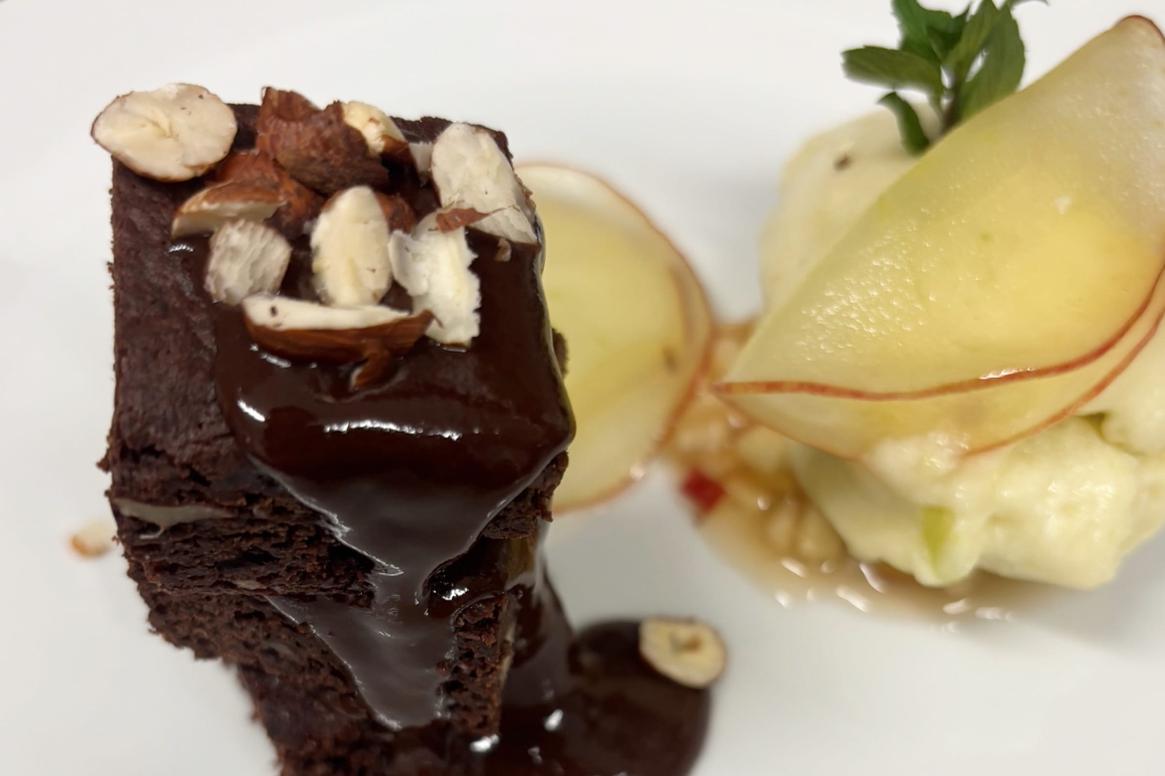 Schokoladenkuchen mit Schokoladensauce und gehackten Nüssen neben Birnenscheiben auf weißem Teller.