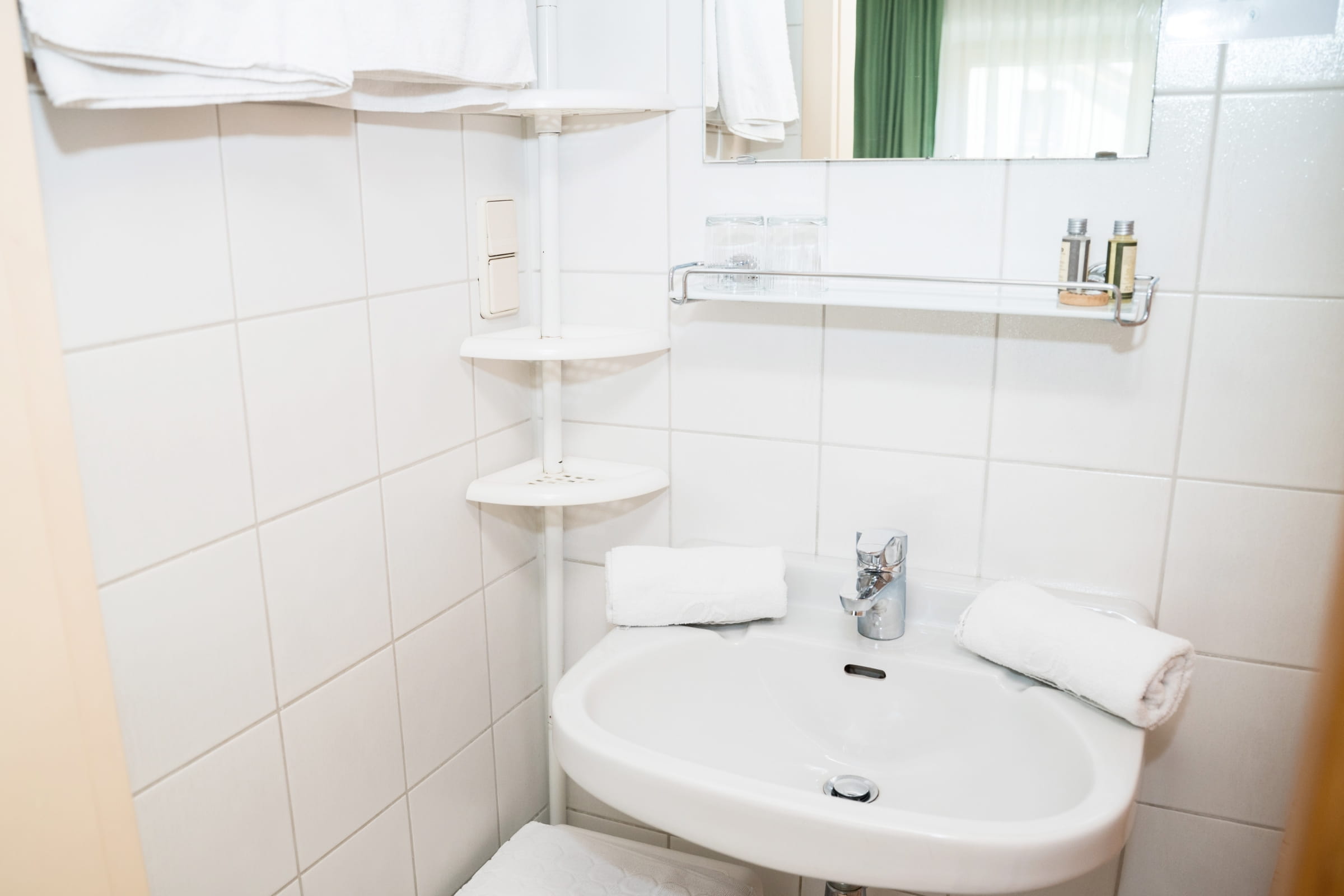 Badezimmer mit weißem Waschbecken, Handtüchern, Wandregal und weißen Fliesen.