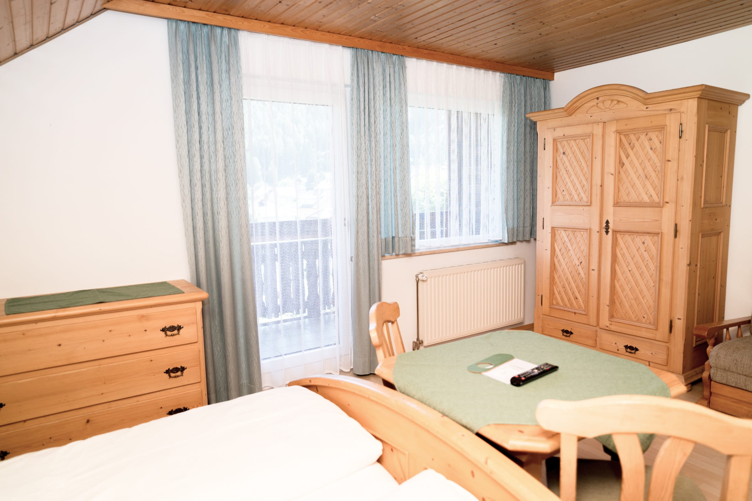 Zimmer mit Holzmöbeln, Bett, Tisch mit Stühlen, Kommode und Balkon mit Vorhängen.