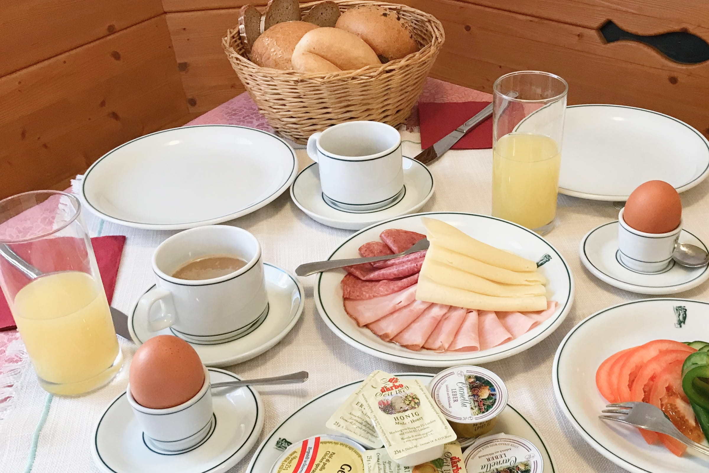 Frühstückstisch mit Brötchenkorb, Kaffee, Orangensaft, gekochten Eiern, Aufschnitt, Käse, Butter und Tomatenscheiben in unserem Gasthof.