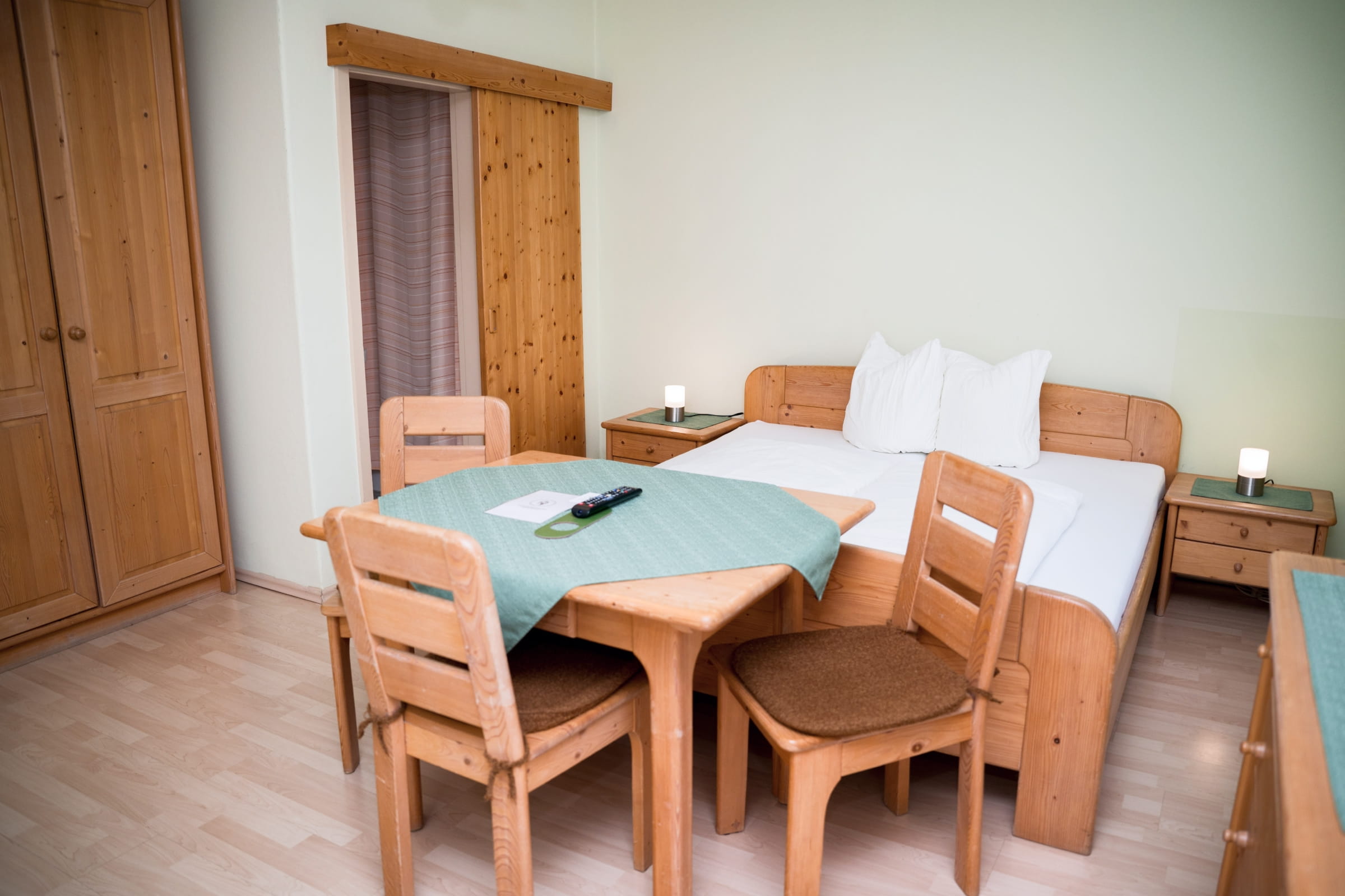 Zimmer mit Holzmöbeln, Doppelbett, Tisch mit drei Stühlen und zwei Nachttischen mit Lampen.