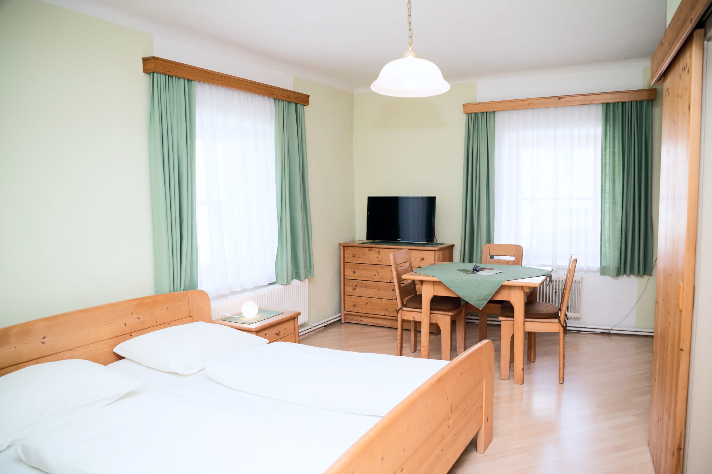 Hell eingerichtetes Gästezimmer mit Doppelbett, Holzmöbeln, Esstisch mit Stühlen und Fernseher vor zwei Fenstern mit grünen Vorhängen.